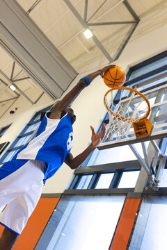 Jumping african american male in blue jersey dunking basketball wearing gym with glass backboard