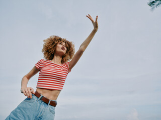 Female dancer outdoors with curly hair, wearing a striped crop top and denim jeans, reaching upward...