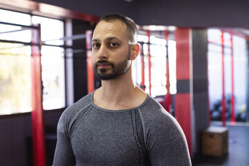 Indian man in gray sports shirt standing in gym strength area by rig, wooden box