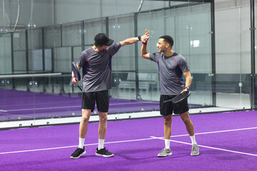 Diverse male teammates celebrating by high-fiving at purple padel court with rackets, wristbands