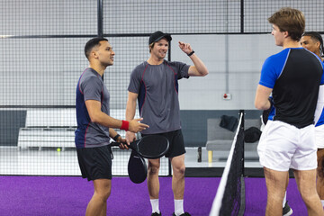 Diverse male teammates standing at net on purple turf padel court and chatting with rackets