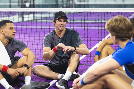Diverse male teammates resting on purple padel court smiling, chatting with towels and racket