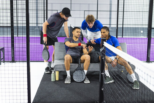 Diverse male friends in sportswear with padel rackets, watching smartphone on purple padel court