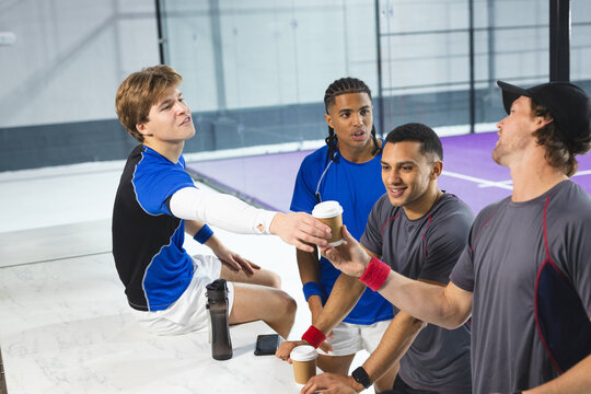 Diverse male teammates in sports gear sitting on squash court passing coffee cup next to bottle