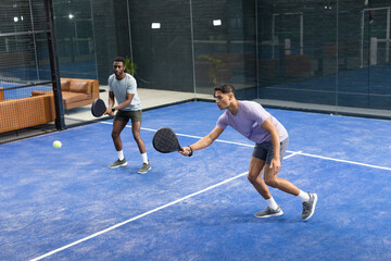 Diverse male teammates hitting yellow padel ball with rackets on padel court next to brown sofas