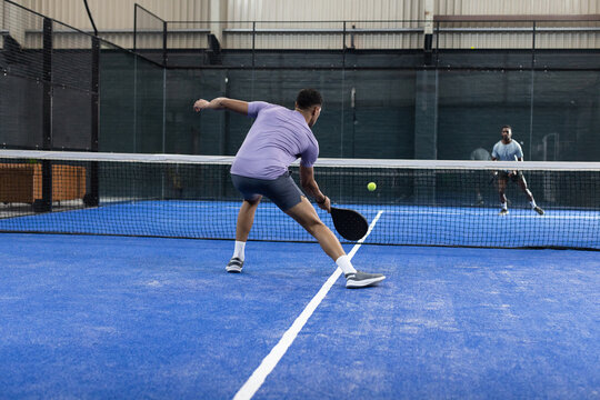 Diverse men rallying on padel court hitting lime green ball with paddle next to net metal fencing