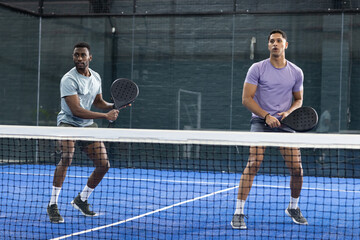 Diverse male teammates standing at net on blue turf padel court, holding rackets, returning shot