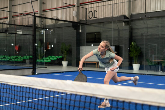 Woman lunging by net on blue padel court with black racket wearing green top, copy space