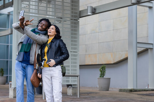 Diverse friends posing for selfie in outdoor urban plaza with smartphone, skateboard, copy space