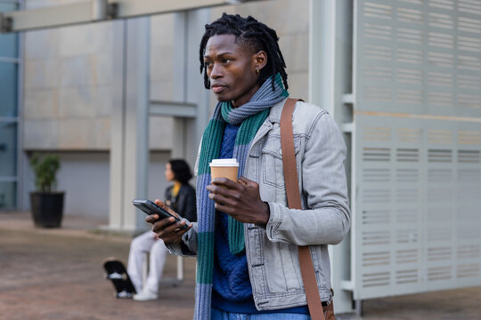 African american man in denim jacket holding coffee cup, using smartphone in plaza