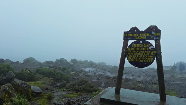 Panning shot shows Baranco Camp sign on a foggy alpine plateau at Mount Kilimanjaro, with a yellow tent and rugged rock wall in the background.