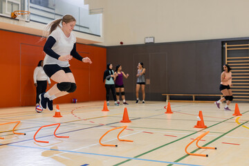 Diverse female teammates performing jumping drills over orange hurdles, cones inside gymnasium