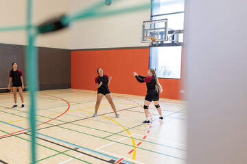 Diverse female teammates positioning under descending volleyball on gym court, green net
