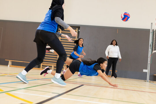 Diverse female volleyball team lunging and diving on gym court under net chasing volleyball