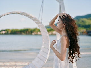 A serene portrait of a young woman standing on a sunlit seaside beach, wearing a white dress, with...