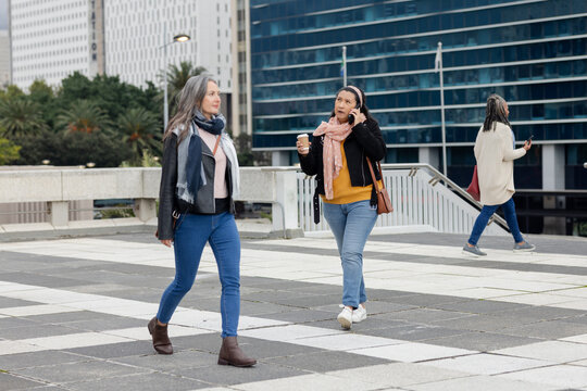 Senior african american and mature white women walking across plaza holding phone, coffee cup