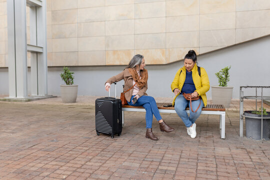 Female travelers sitting on wooden bench at urban plaza holding suitcase, rummaging through bag