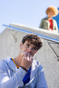 Man sitting on outdoor stairs, biting sandwich, wearing earbuds and hoodie by metal handrail
