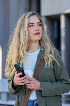 Woman standing outside plaza wearing bag strap with coffee cup smartphone looking right