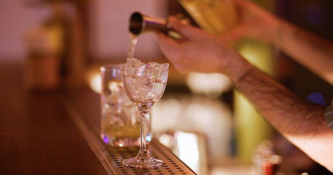 Cocktail making. Using jigger. Barman hand pouring yellow liquid for chilled alcoholic drink from metal measuring cup to glass ice cubes.