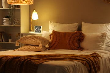 Comfortable bed with blanket, pillows and glowing lamp on bedside table at night