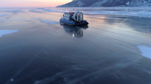 Winter Adventure travel concept. Drone follows fast rescue hovercraft hivus on cracked Ice lake Baikal. Breathtaking Siberian sunset. Tourism destination. Air Cushion Vehicle Racing. Cracks, mountains