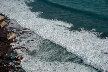 High angle drone shot of a massive white wave breaking on a rugged shoreline. A powerful and abstract view of raw nature's energy.
