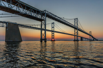 Chesapeake Bay Bridge at yellow and orange sunrise.  Wide angle from beneath bridge with reflection in calm water.