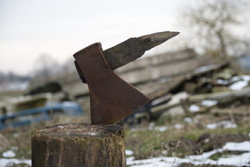 An antique axe with a rusty blade sits in a wooden stump against a snowy, blured background.