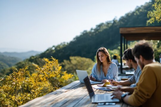 Young professionals working together outdoors on laptops and documents at a wooden table, surrounded by greenery and mountains in a modern remote work environment
