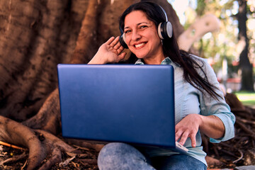 Beautiful adult Latina professional woman working on her laptop, wearing headphones and listening...