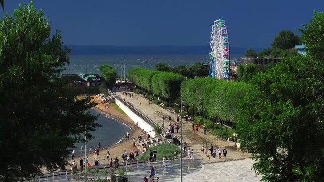 Vibrant promenade at Sports Harbor embankment in Vladivostok, Russia, along Amur Bay with Ferris wheel, sandy beach, strolling crowds, lush green hedges, and waterfront cafes.
