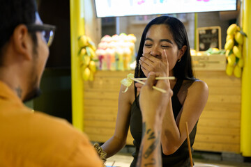 Man feeding girlfriend with chopsticks at asian food stall, couple sharing a funny moment