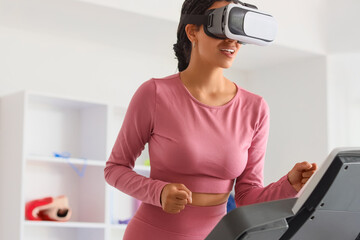 Young African-American woman in VR glasses running on treadmill at gym