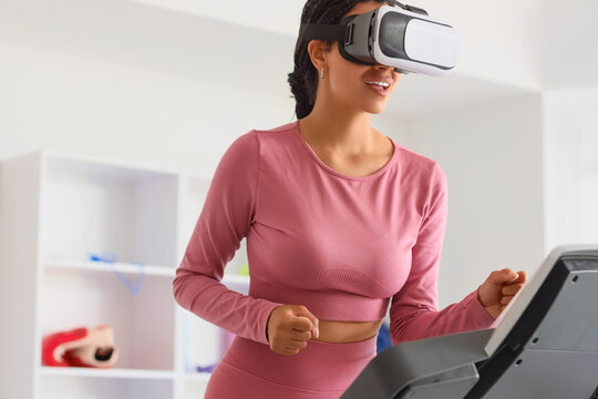 Young African-American woman in VR glasses running on treadmill at gym