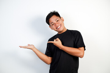 A young Asian man smiling cheerfully, feeling happy and pointing to the side, showing object in...
