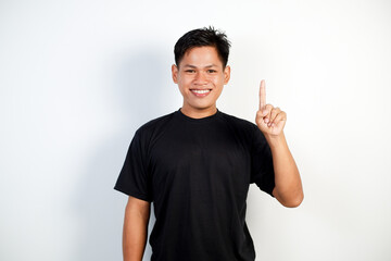 A Cheerful Young Asian Man Wearing a Black Shirt Pointing Upward With a Smile on a White Background.