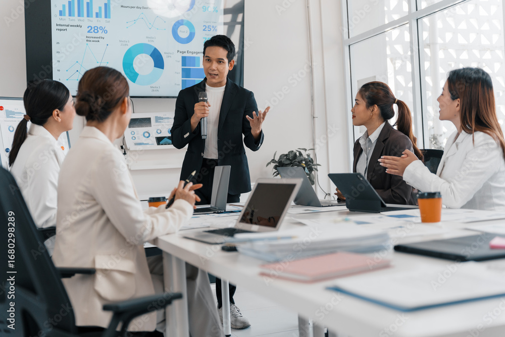 Poster businessman using microphone while giving presentation to colleagues in boardroom, in front of chart