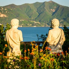 Two ancient statues overlook a serene blue sea and lush green mountains.