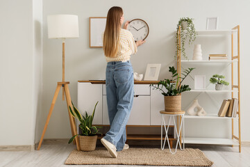 Young woman hanging clock on light wall at home, back view