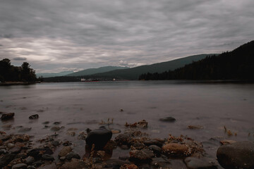 A tranquil long-exposure shot of a rocky shoreline leading to calm, misty water. Distant mountains and trees frame the horizon under an overcast sky, with subtle hints of urban development