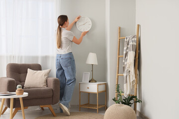Young woman hanging clock on light wall in living room, back view