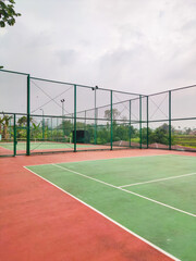 Empty tennis court with green and red surface, surrounded by tall wire fencing under an overcast sky, creating a quiet atmosphere for practice, competition, or leisure play in a peaceful outdoor setti