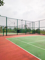 Empty tennis court with green and red surface, surrounded by tall wire fencing under an overcast sky, creating a quiet atmosphere for practice, competition, or leisure play in a peaceful outdoor setti