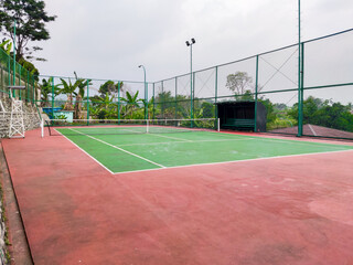 Fototapeta premium Empty tennis court with green and red surface, surrounded by tall wire fencing under an overcast sky, creating a quiet atmosphere for practice, competition, or leisure play in a peaceful outdoor setti