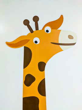 Playful cartoon giraffe wall decal features bold yellow and brown patterns with big expressive eyes, painted on a plain white wall in a whimsical, child-friendly style.