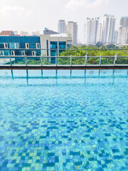 Rooftop swimming pool with mosaic tiles reflecting sunlight. In the background, tall city buildings rise above greenery, blending urban lifestyle with relaxation.