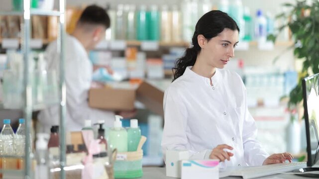 Near counter with computer is pharmacists workplace. Girl employee participates in online testing and learning, reads incoming emails. Electronic database of drugs. Colleague work in background
