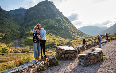 Mother and teenage son standing on stone wall in Glencoe Highlands pointing at the sky and enjoying...