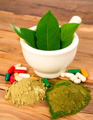 Fresh green leaves rest in a white mortar, alongside herbal powder and assorted pills, on a light-brown wooden surface.